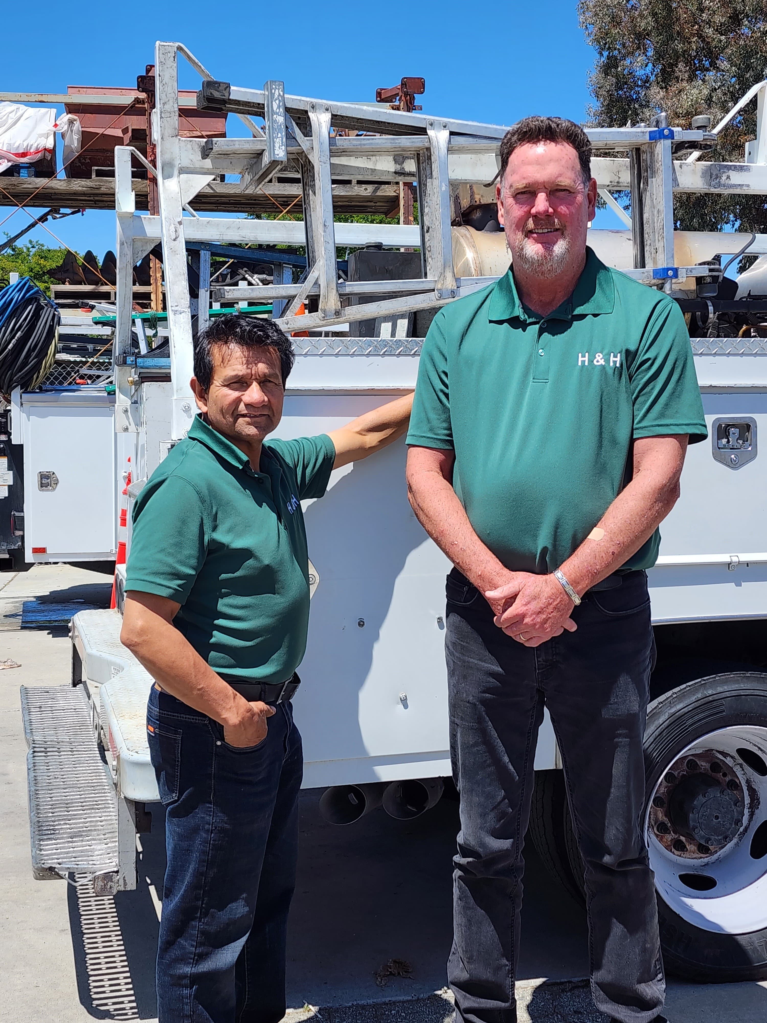 Two men in green H&H polo shirts stand beside a white utility service truck.